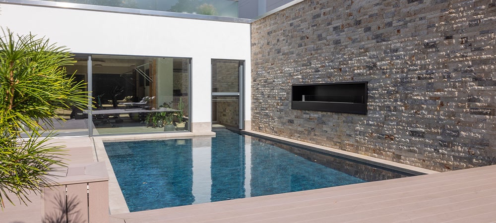 Moderner Pool im Garten, umgeben von Holzdielen und Steinwand, reflektiert Glasfront des Hauses.