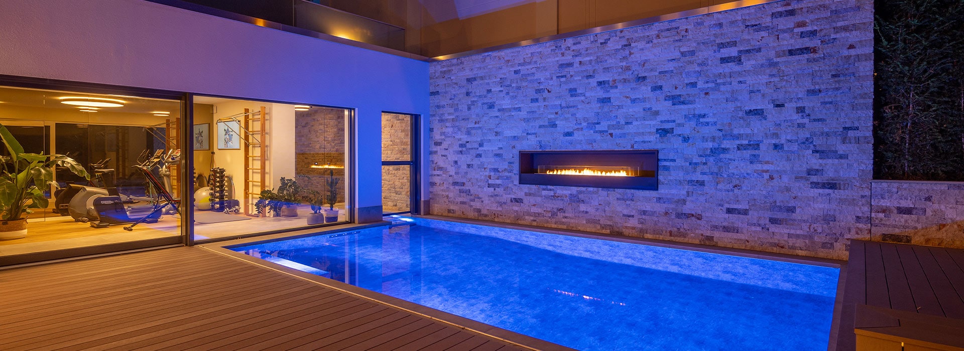 Luxuriöser Innenhof mit blau erleuchtetem Pool, Kamin an Steinwand, Fitnessraum links.