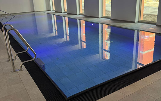 Innenpool mit blauer Beleuchtung, Metallgeländer links, große Fenster, helles Design.